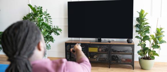 Seated Black man, shown from behind, points a remote at a blank TV screen.